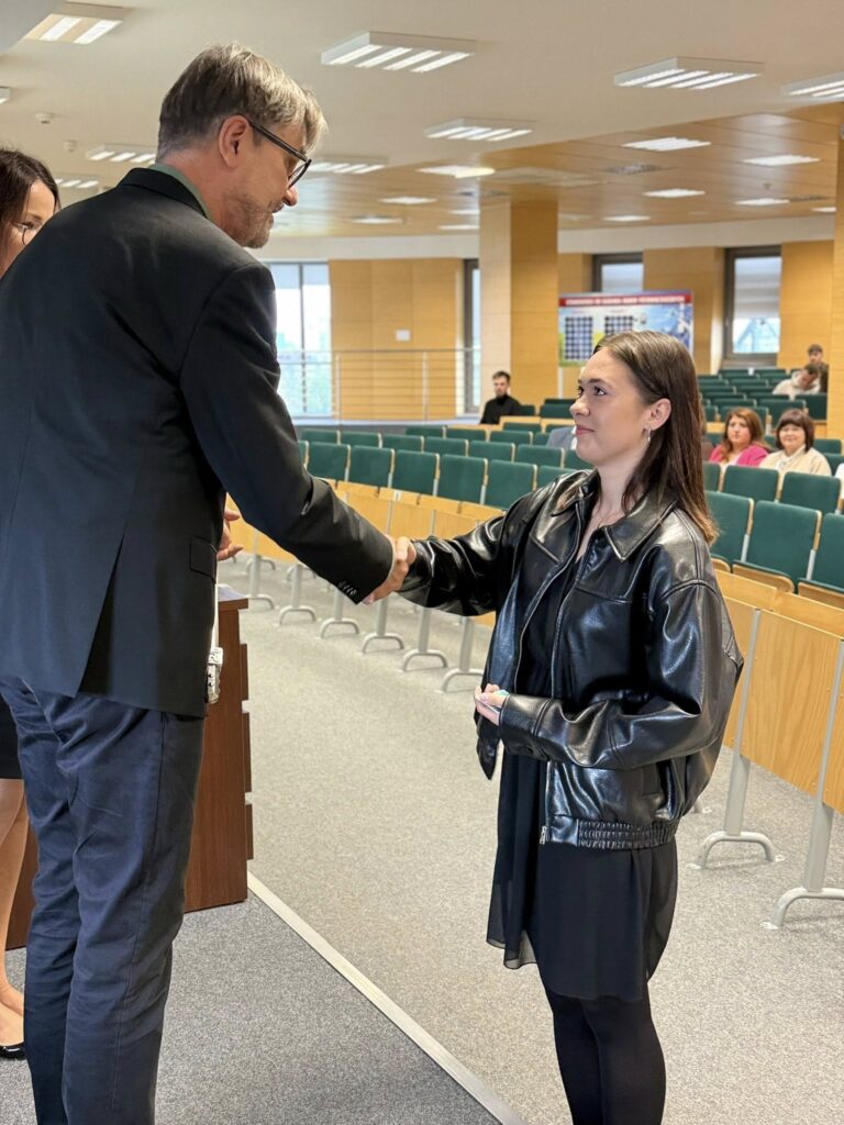 Mężczyzna w garniturze i kobieta w skórzanej kurtce podają sobie ręce. Kontekst: sala wykładowa.