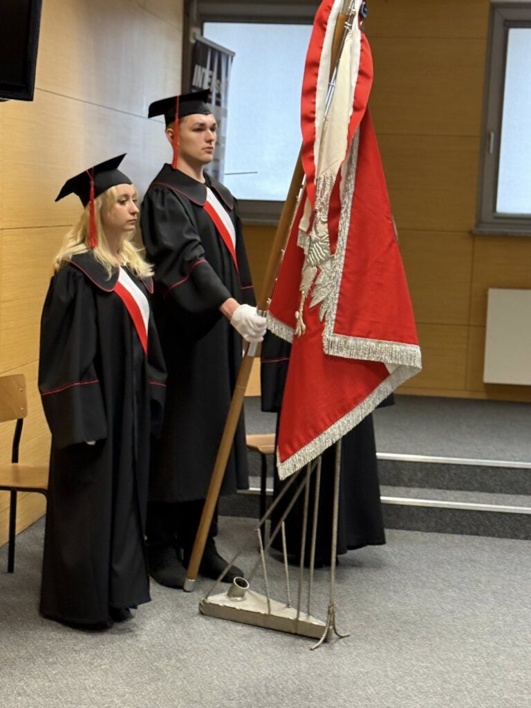 Dwoje studentów w togach akademickich stoi obok sztandaru. Kluczowe obiekty: togi, birety, sztandar.