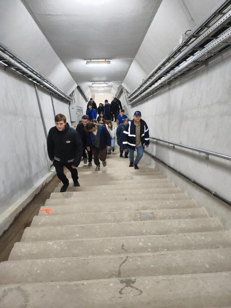 Grupa osób schodzi po betonowych schodach w długim korytarzu. Widać kable i oświetlenie.
