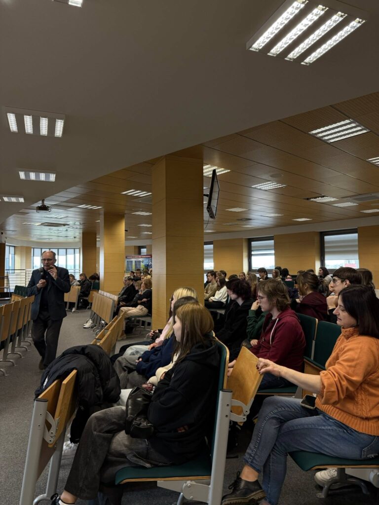 Mężczyzna w garniturze przemawia do grupy studentów siedzących na widowni. Kontekst: wykład lub prezentacja.