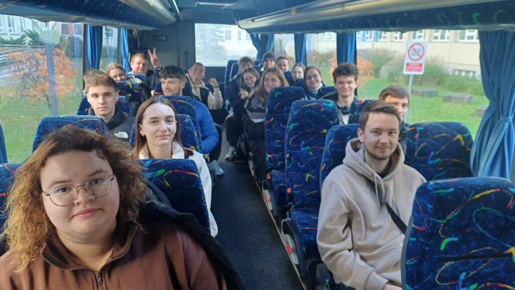 Grupa młodych ludzi siedzi w autobusie. Widać siedzenia, okna i drzewa za oknem.