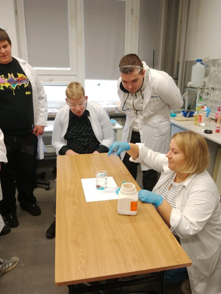 Grupa osób w fartuchach laboratoryjnych przy stole z naczyniami i odczynnikami. Kontekst: lekcja chemii.