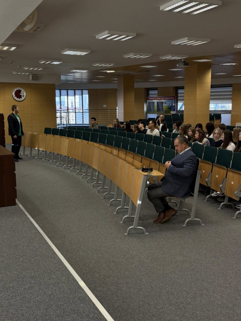 Wykład w auli. Mężczyzna w garniturze siedzi, inny stoi. Studenci na widowni.