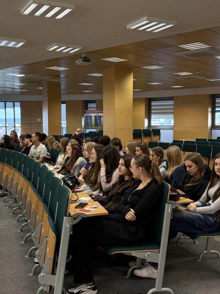 Grupa studentów siedzi na wykładzie w sali seminaryjnej. Kluczowe obiekty: studenci, krzesła, stoły, projektor.