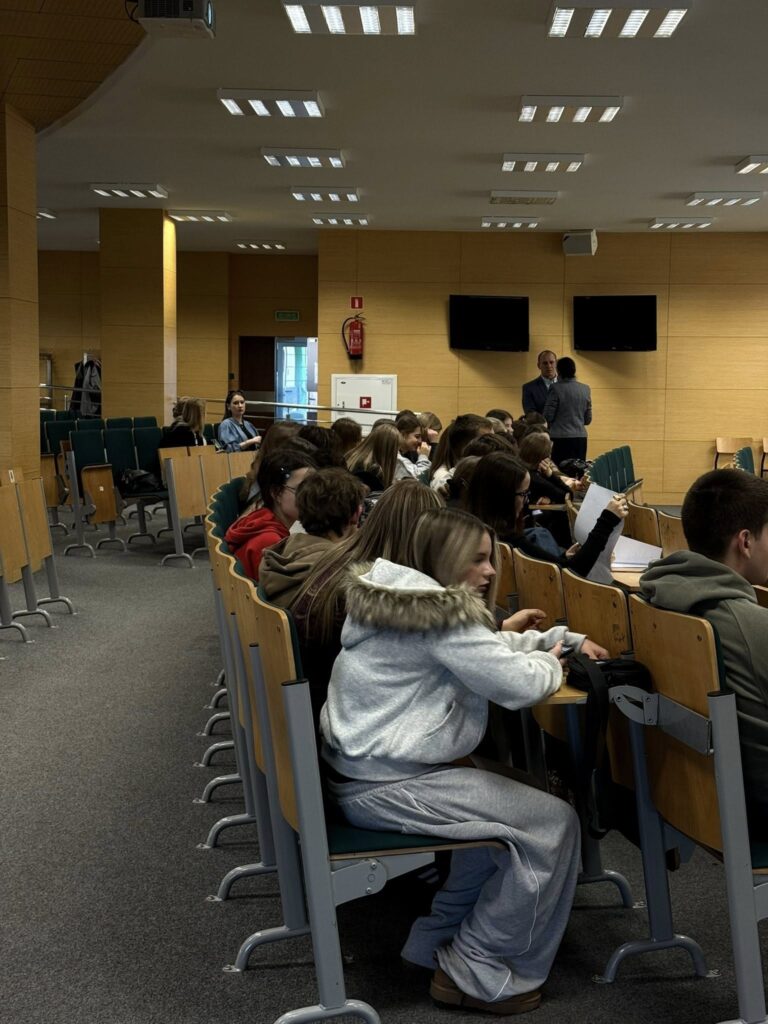Widownia z siedzącymi uczniami w sali wykładowej. W tle dwaj mężczyźni stojący przed ekranami.