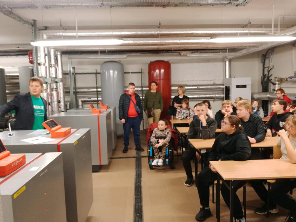 Grupa osób, w tym młodzież, ogląda instalację grzewczą. W pomieszczeniu znajdują się kotły, rury i grzejniki.