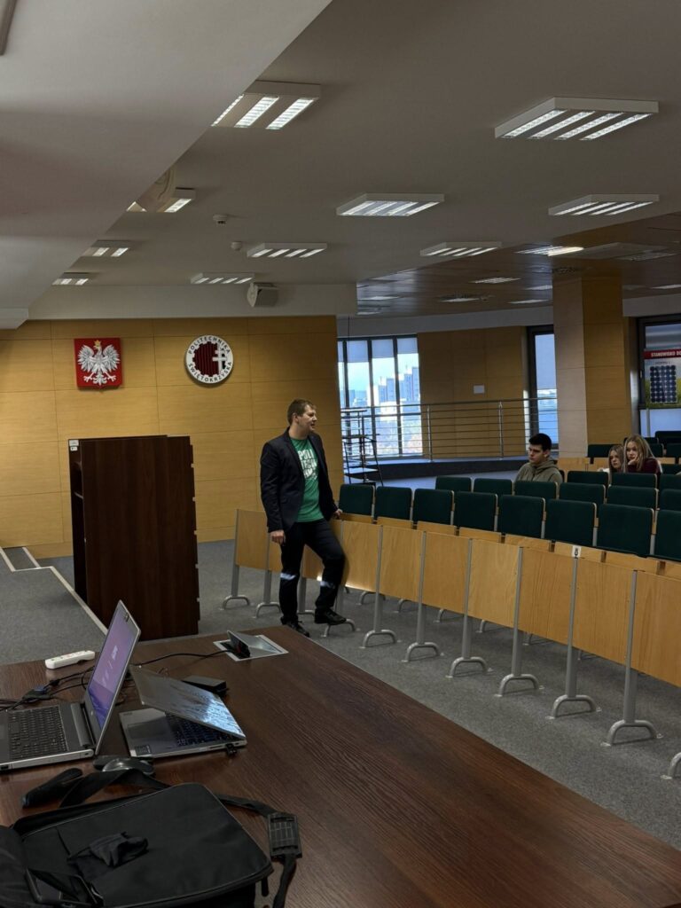 Mężczyzna w garniturze stoi w sali wykładowej. Widać ławki, laptopy i godła.