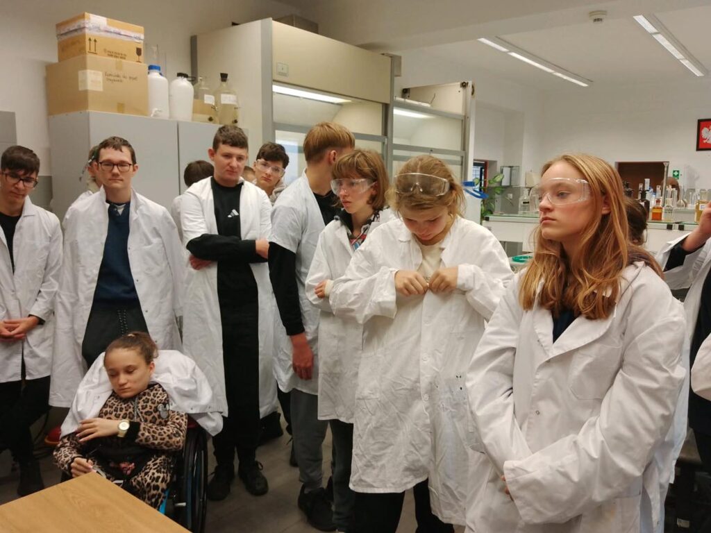 Grupa uczniów w fartuchach laboratoryjnych stoi w sali. Wszyscy noszą okulary ochronne.