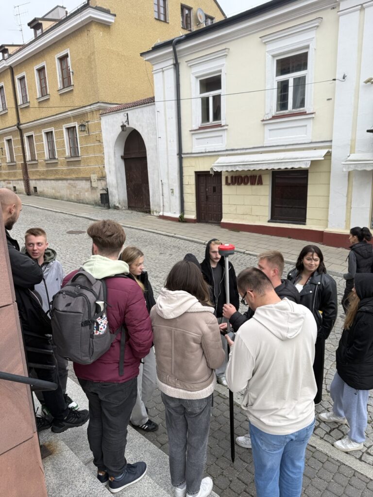 Grupa młodych ludzi stoi na ulicy przed budynkami. W tle widoczny szyld "LUDOWA".