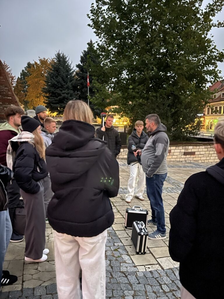 Grupa ludzi stoi na zewnątrz, słuchając mężczyzny. W tle drzewa i budynki.
