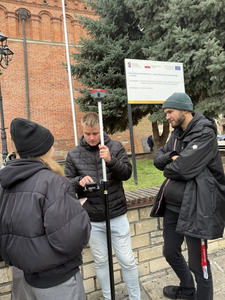 Trzy osoby przy geodezyjnym sprzęcie pomiarowym. W tle tablica informacyjna o rewitalizacji.