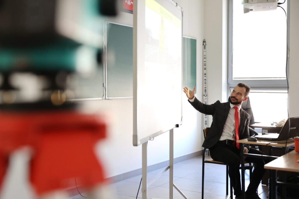 Mężczyzna w garniturze i czerwonym krawacie gestykuluje przy tablicy. W tle widać projektor i okno.