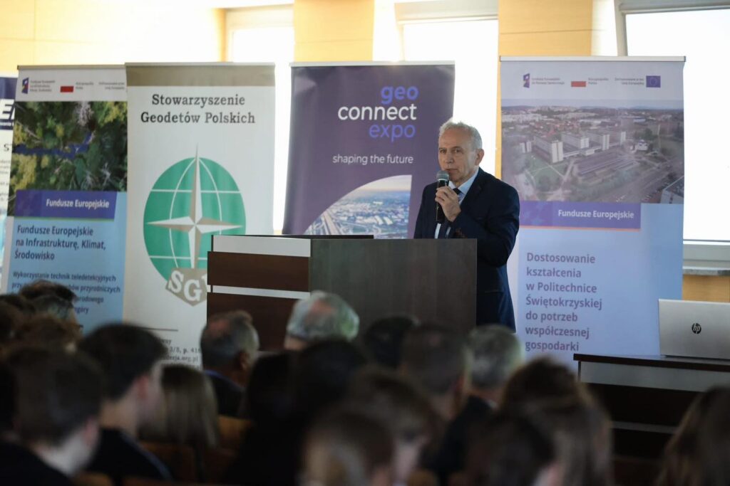 Mężczyzna przemawia na tle banerów "geo connect expo", "Stowarzyszenie Geodetów Polskich" i "Fundusze Europejskie".