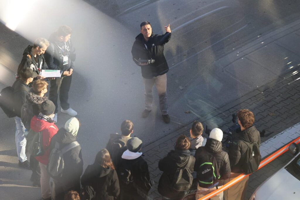 Mężczyzna z dronem przemawia do grupy osób. Kluczowe obiekty: mężczyzna, dron, grupa ludzi, droga, chodnik.