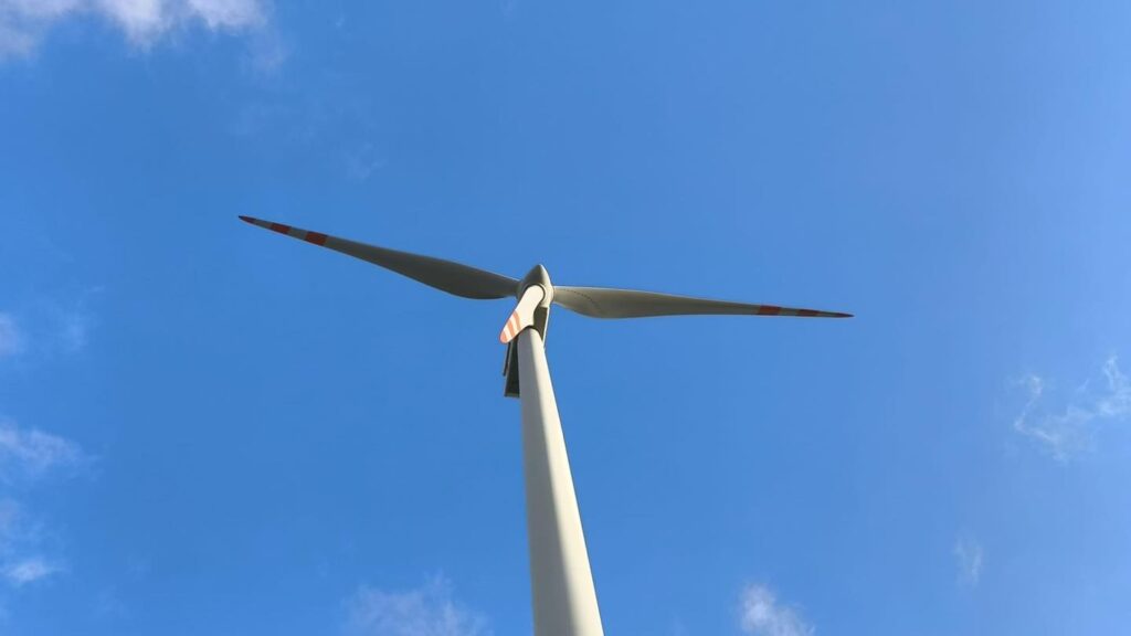 Wiatrak z białym masztem i łopatami na tle błękitnego nieba z chmurami. Kontekst: energia odnawialna.