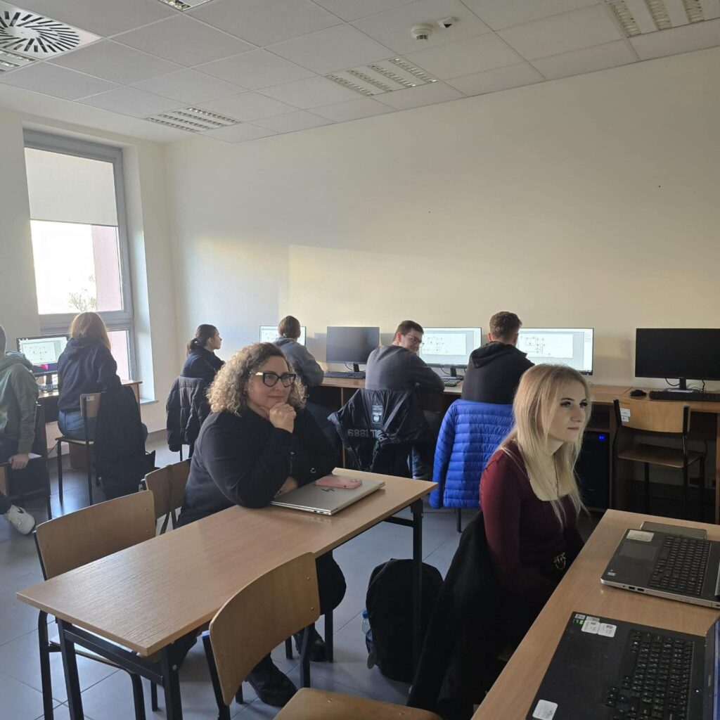 Osoby siedzą przy biurkach w sali lekcyjnej z komputerami. Widać laptopy, monitory i krzesła.