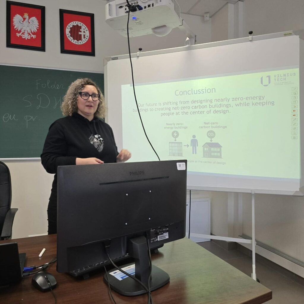 Kobieta prezentuje na ekranie koncepcję budynków zeroemisyjnych. W tle tablica z notatkami i godła.