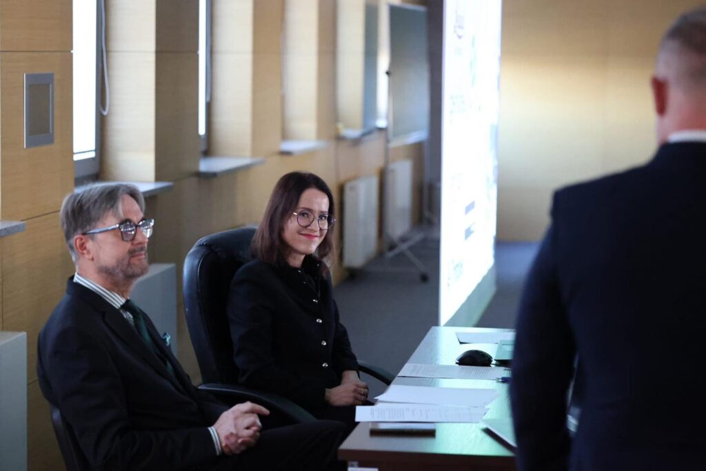 Mężczyzna i kobieta siedzą przy stole w sali konferencyjnej. W tle widać ekran.