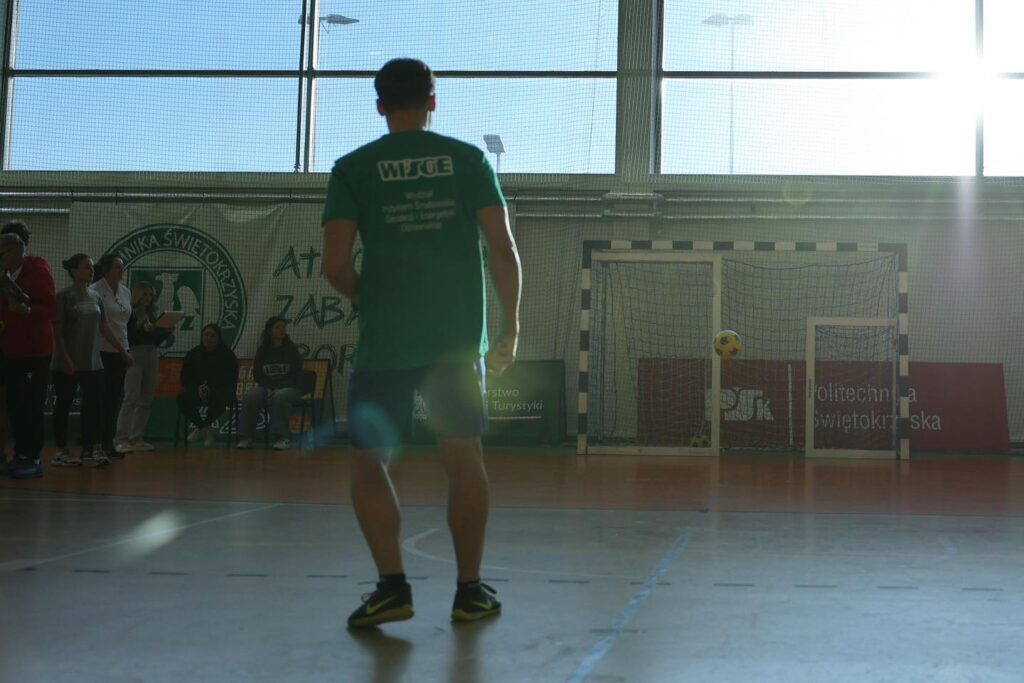 Mężczyzna w zielonej koszulce stoi na hali sportowej. W tle bramka, piłka i widzowie.
