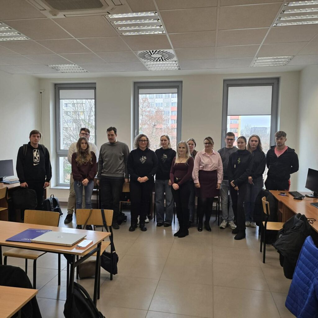 Grupa osób stoi w sali lekcyjnej. Widać okna, biurka i krzesła.