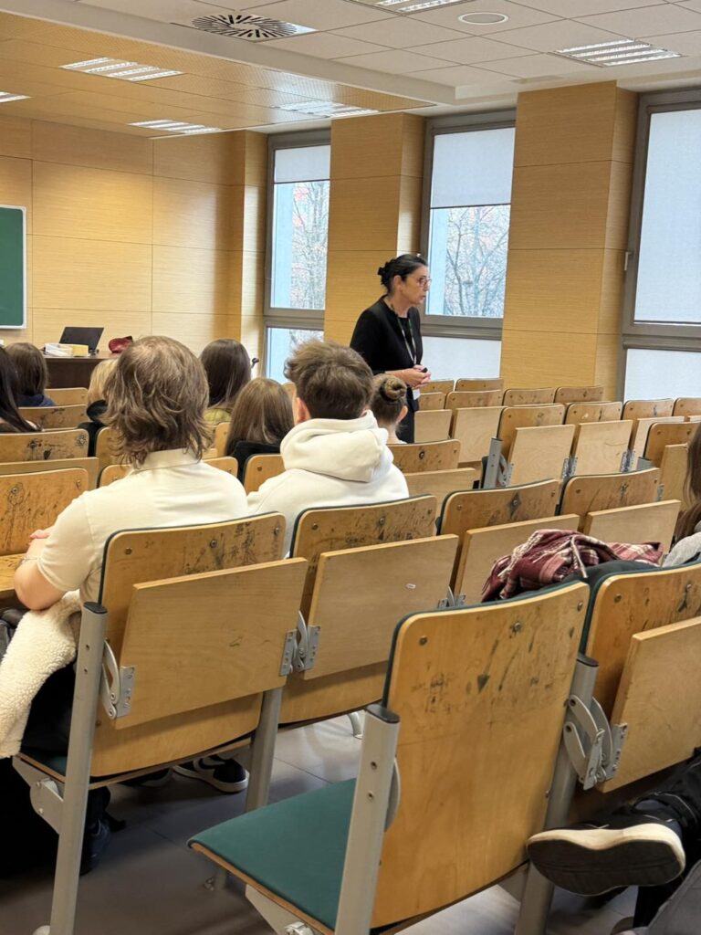 Kobieta prowadzi wykład dla grupy studentów siedzących w sali. Kluczowe obiekty: wykładowca, studenci, ławki, okna.
