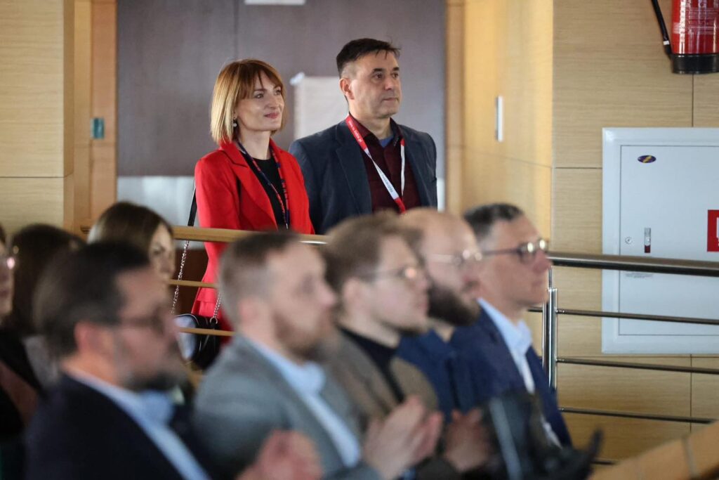 Kobieta w czerwonej marynarce i mężczyzna w granatowej marynarce stoją. W tle widownia i gaśnica.