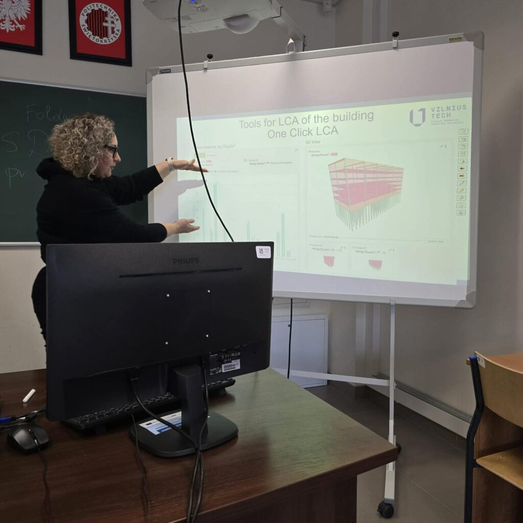 Kobieta wskazuje na ekran z narzędziami do analizy cyklu życia budynku. Kluczowe obiekty: kobieta, ekran, projektor, monitor.