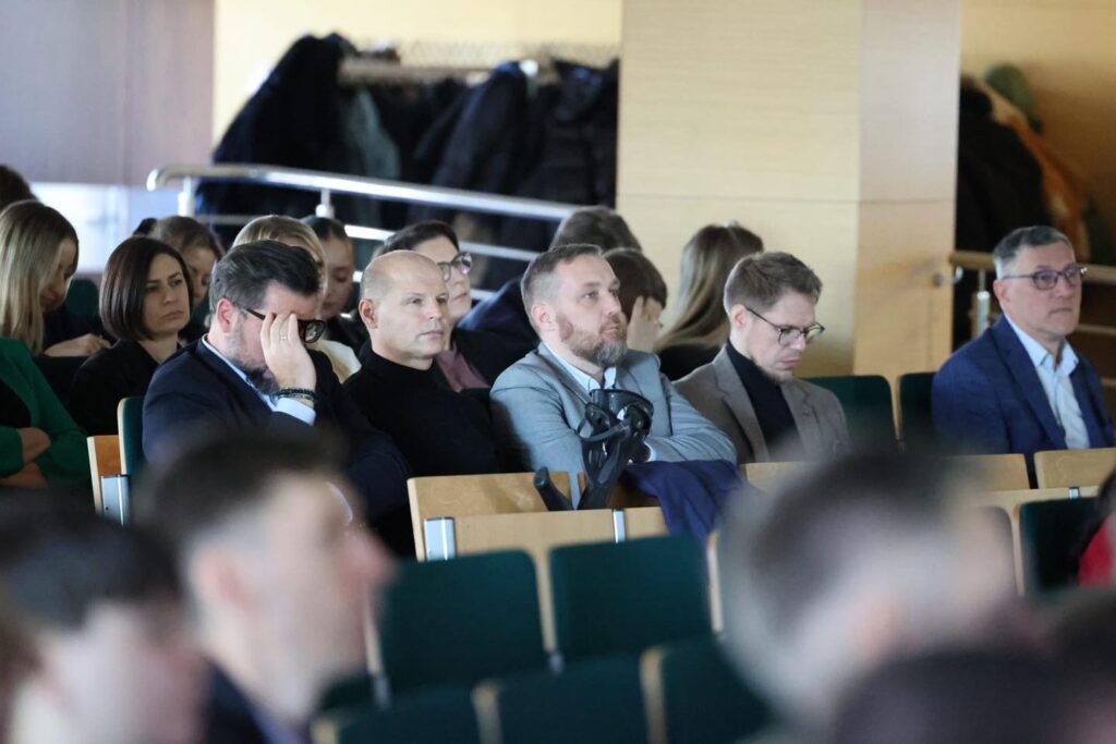 Ludzie siedzą w rzędach na widowni. Widać mężczyzn w garniturach i kobiety. Kontekst to wydarzenie publiczne.