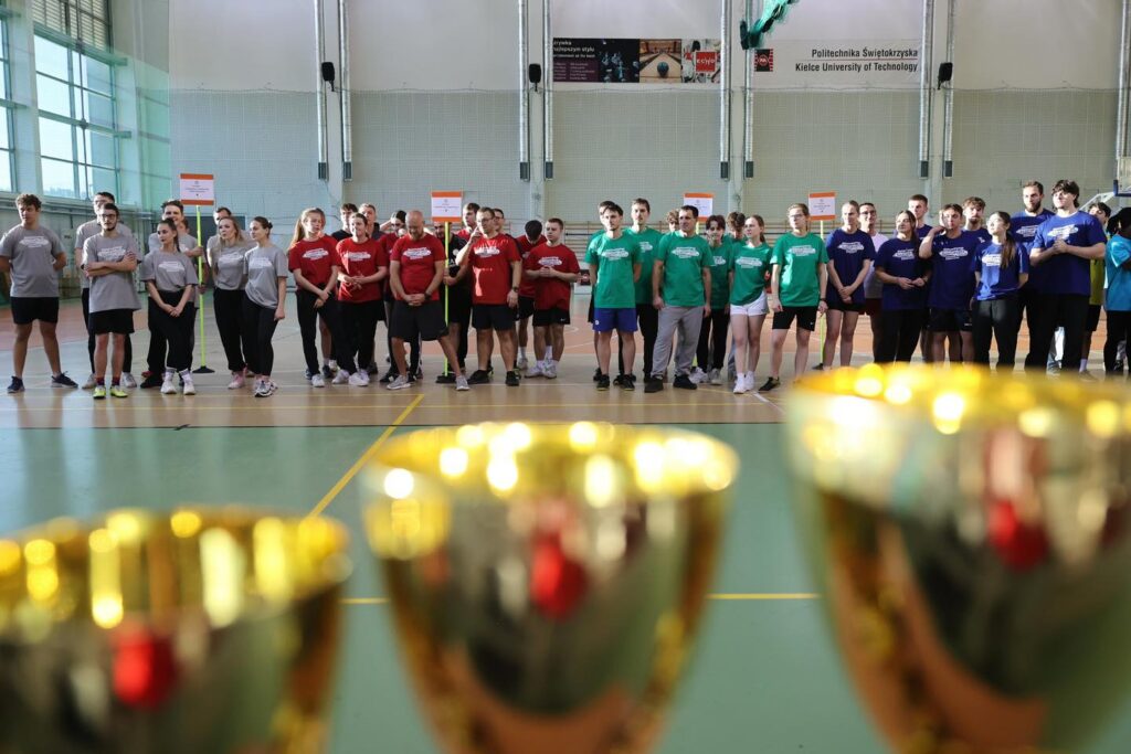 Grupa ludzi w strojach sportowych stoi w hali sportowej. W tle baner Politechniki Świętokrzyskiej.