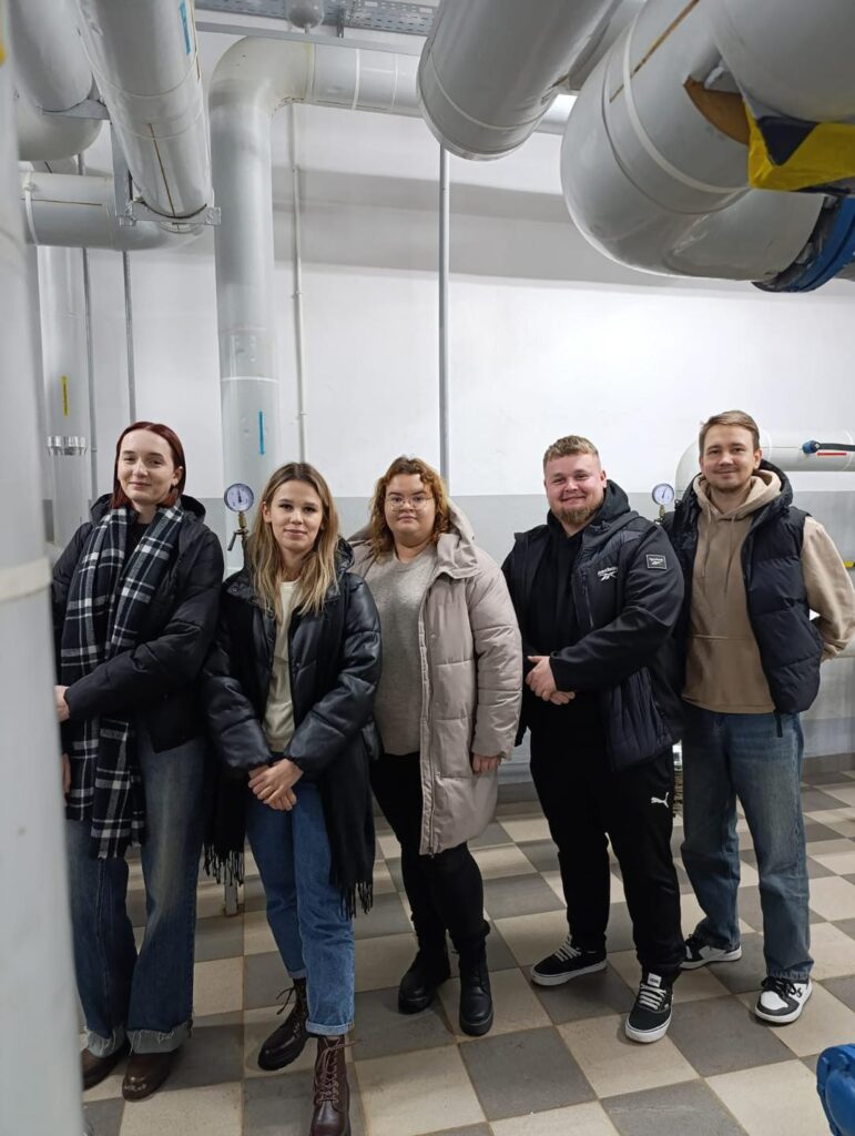 Pięć osób stoi przed dużymi rurami. W pomieszczeniu znajdują się instalacje techniczne.