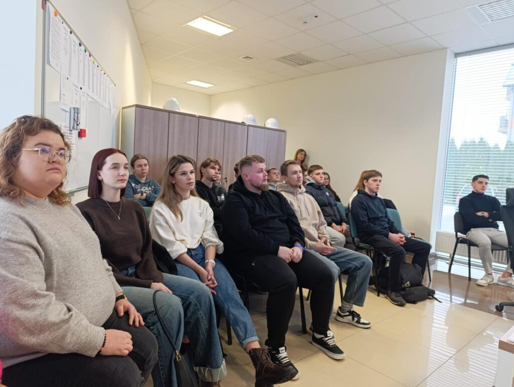 Grupa osób siedzi na krzesłach w sali. Widać tablicę, szafki i okno.