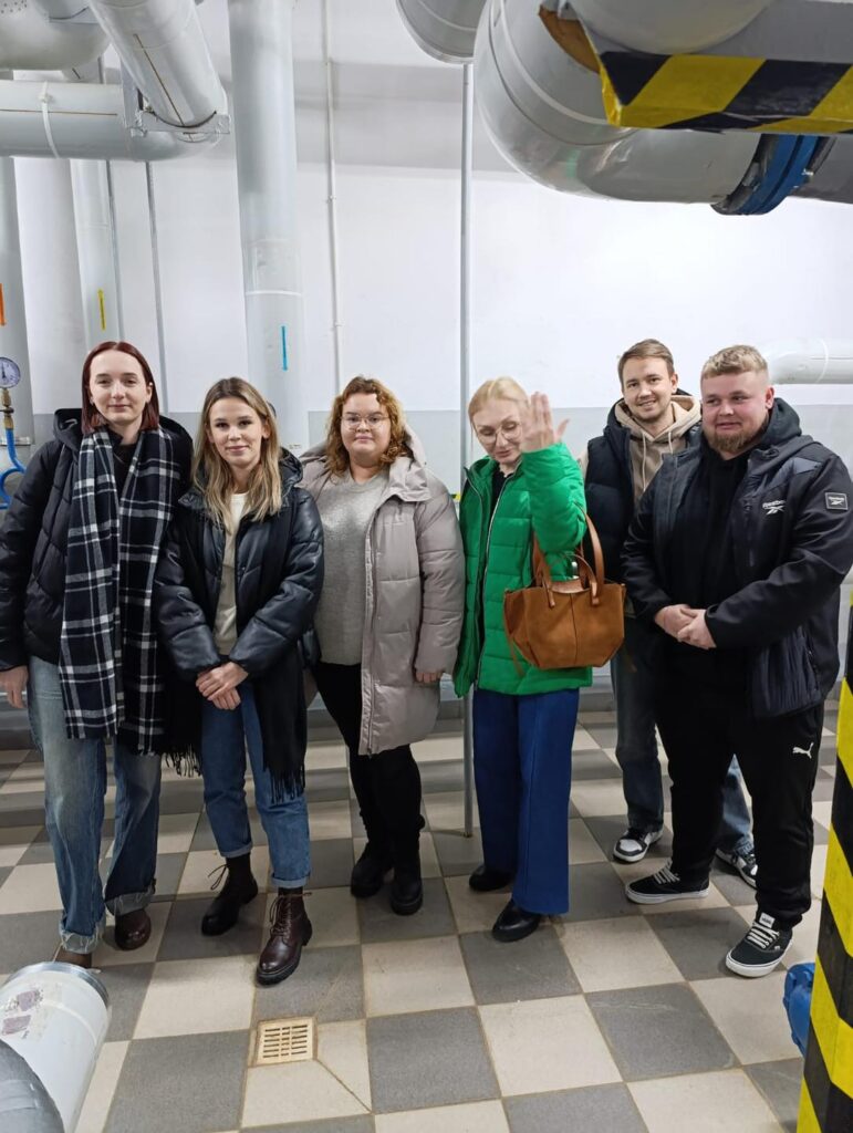Grupa osób stoi w pomieszczeniu z rurami. Widać ludzi, rury, kratkę.