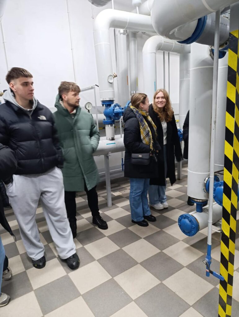 Grupa osób ogląda instalację hydrauliczną z dużymi rurami i zaworami. W pomieszczeniu znajdują się też manometry.