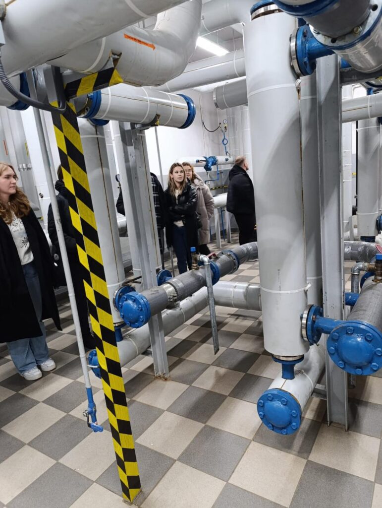 Grupa osób ogląda instalację rurową z zaworami i izolacją. Kluczowe obiekty: rury, zawory, izolacja.