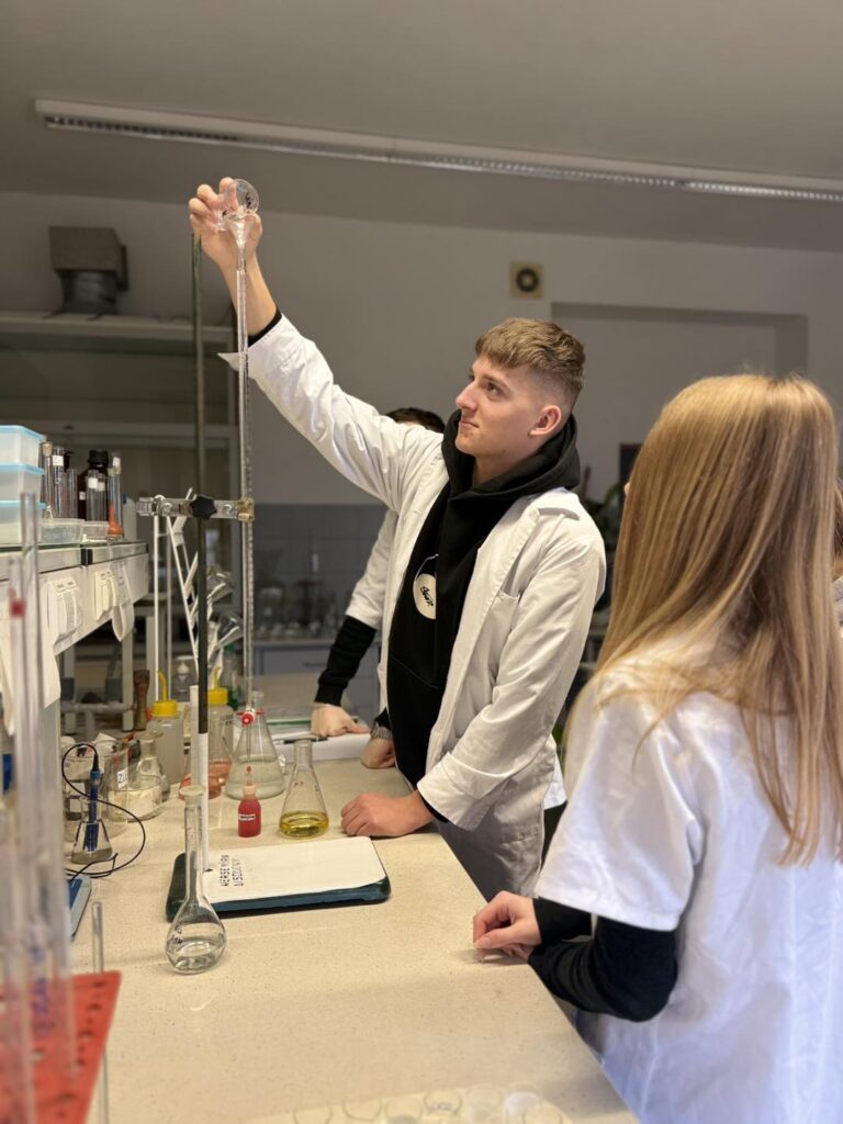 Młody mężczyzna w fartuchu laboratoryjnym używa biurety. W tle widać inne osoby i sprzęt laboratoryjny.