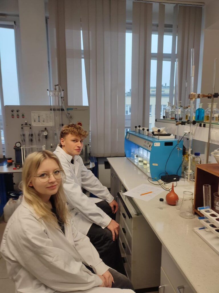 Dwoje studentów w fartuchach laboratoryjnych siedzi przy stole w pracowni.