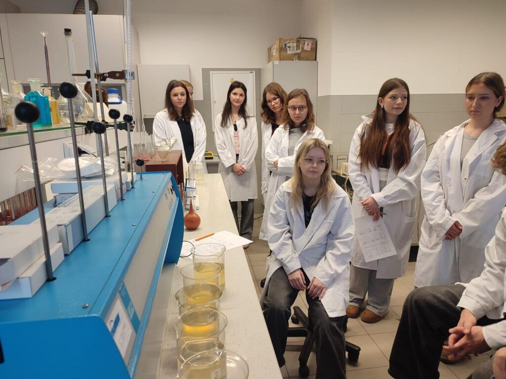 Grupa uczniów w kitlach laboratoryjnych obserwuje eksperyment. W tle sprzęt laboratoryjny, probówki, zlewki z płynem.