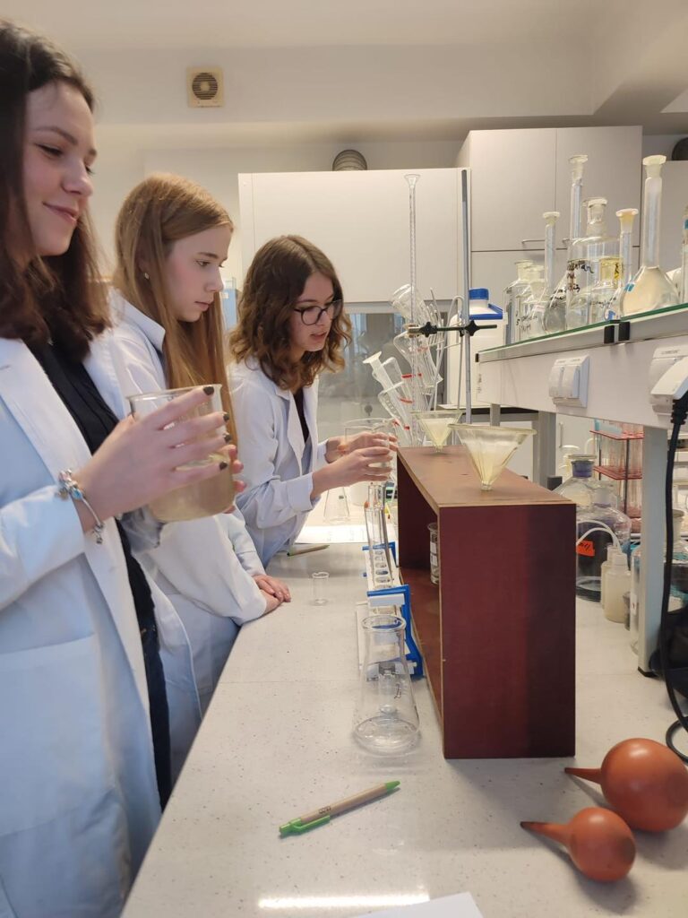 Trzy uczennice w kitlach laboratoryjnych wykonują eksperyment. Kluczowe obiekty: kolby, zlewki, lejki, pipety.