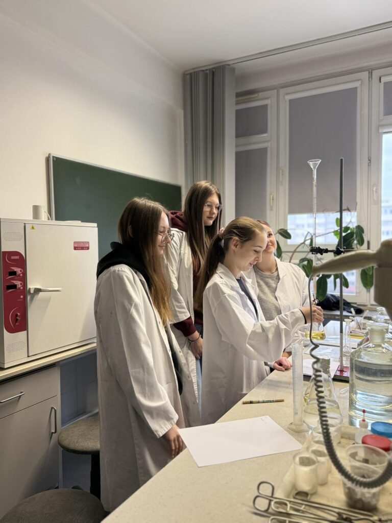 Cztery studentki w kitlach laboratoryjnych wykonują eksperyment. W tle piec laboratoryjny i okno.