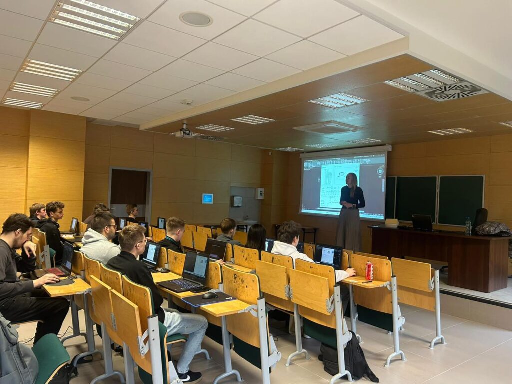 Sala wykładowa z kobietą przy projektorze i studentami przy laptopach.
