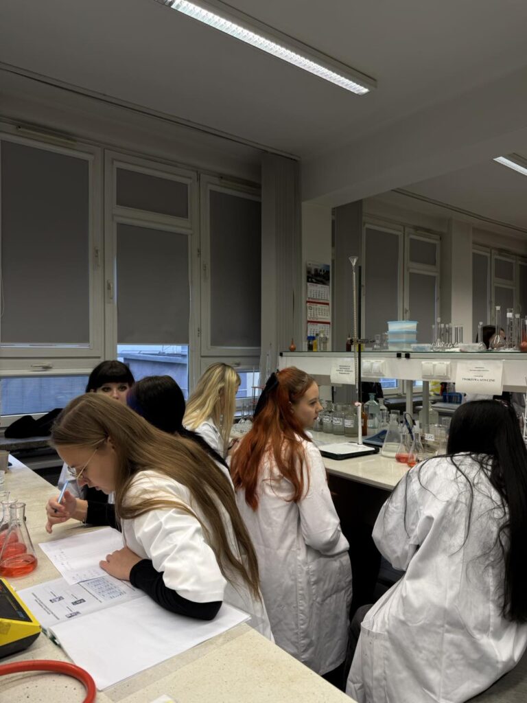 Grupa uczniów w fartuchach laboratoryjnych pracuje przy stole w laboratorium. Widać kolby z płynami i dokumenty.