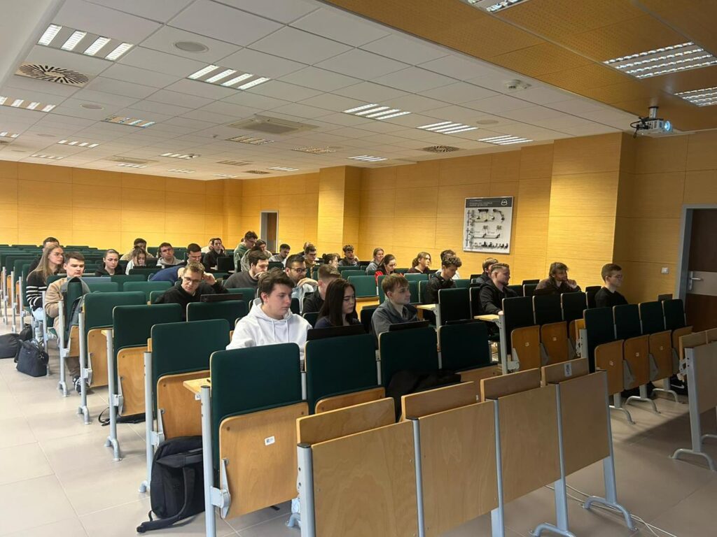 Grupa studentów siedzi w sali wykładowej, patrząc na tablicę. Widać projektor i żółte ściany.