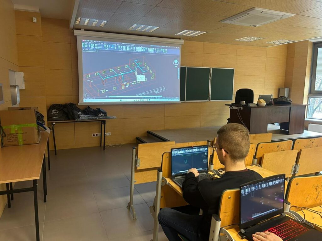 Sala wykładowa z rzutnikiem wyświetlającym projekt 3D. Uczniowie pracują na laptopach.