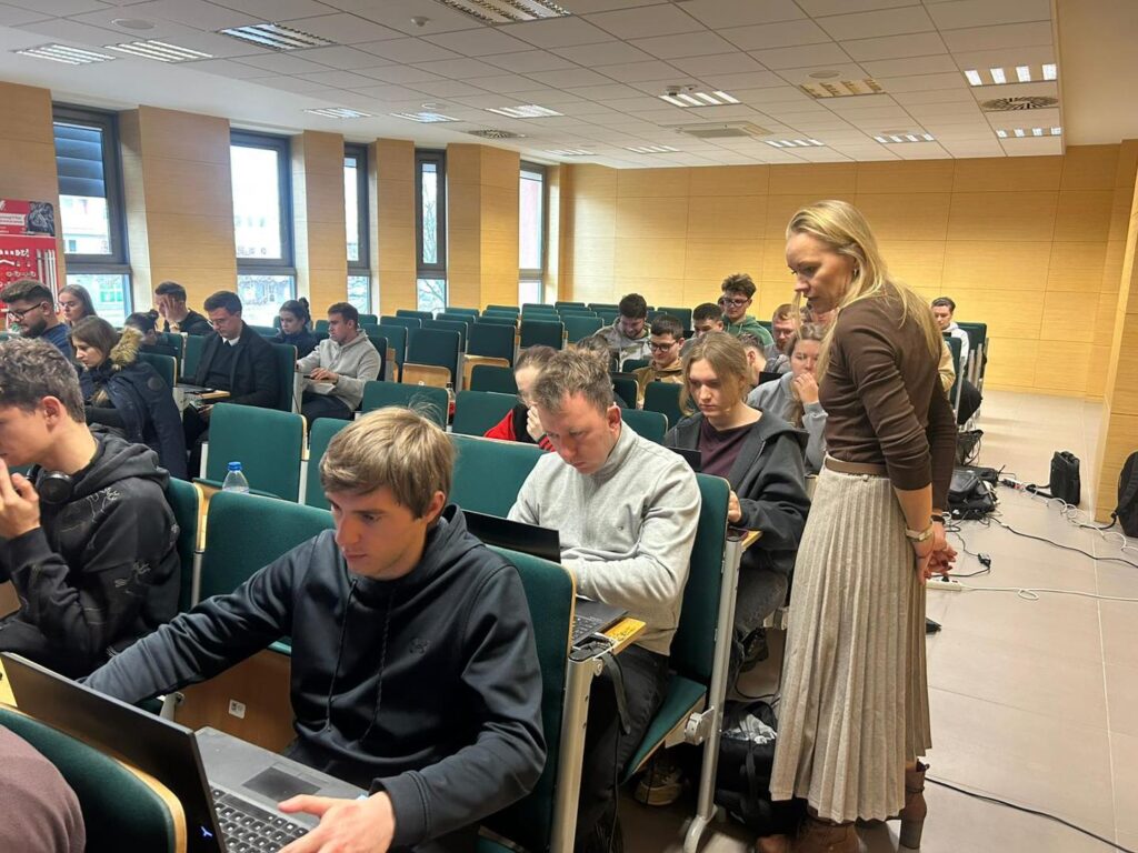 Sala wykładowa z ludźmi przy laptopach. Kobieta stoi, obserwując grupę.