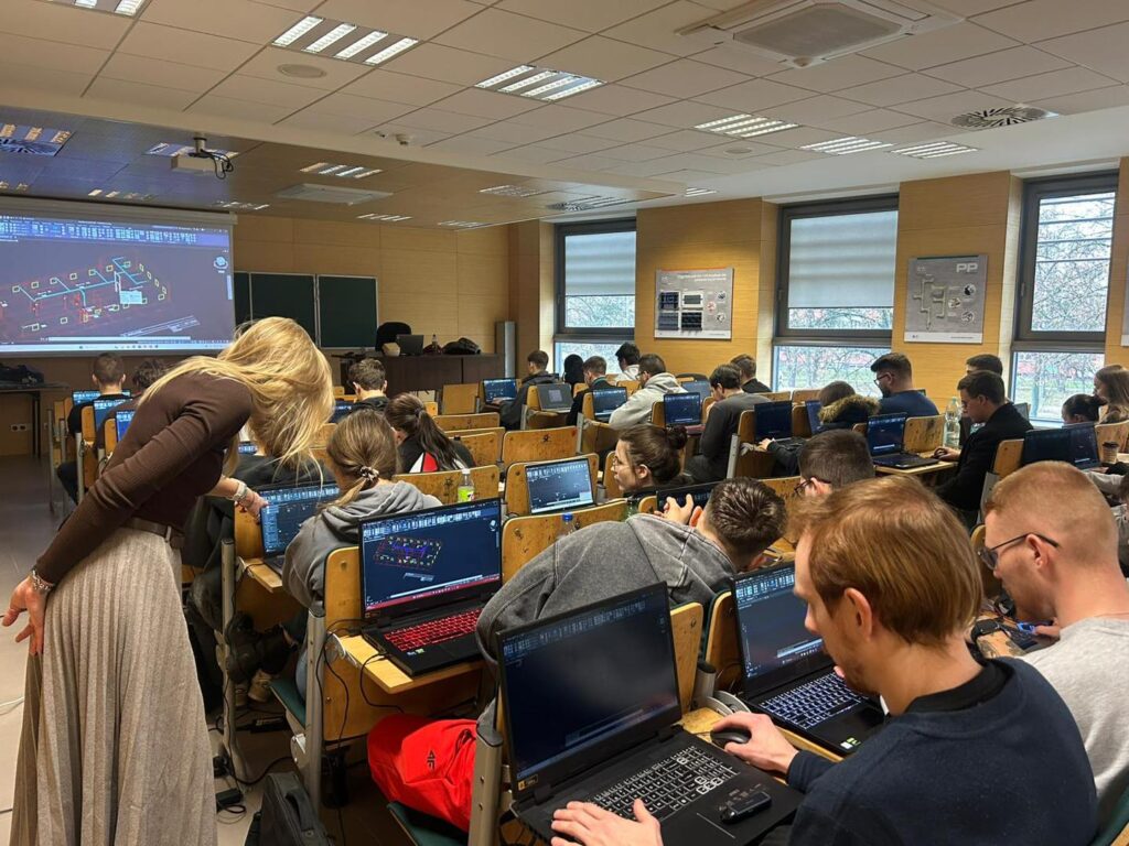 Nauczycielka pomaga studentom przy komputerach w sali wykładowej. Kluczowe obiekty: laptop, projektor, studenci.