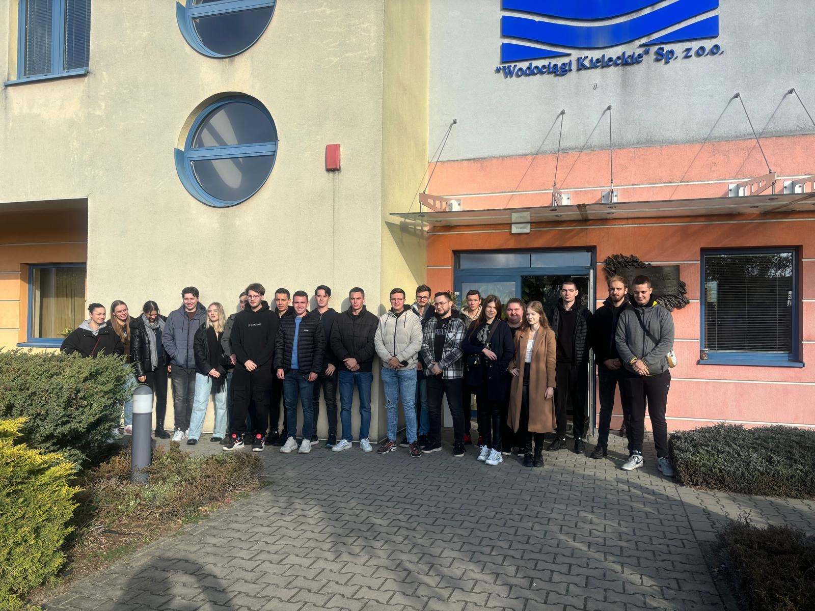 Grupa młodych ludzi stoi przed budynkiem z napisem "Wodociągi Kieleckie". Kluczowe obiekty: budynek, grupa ludzi, napis.