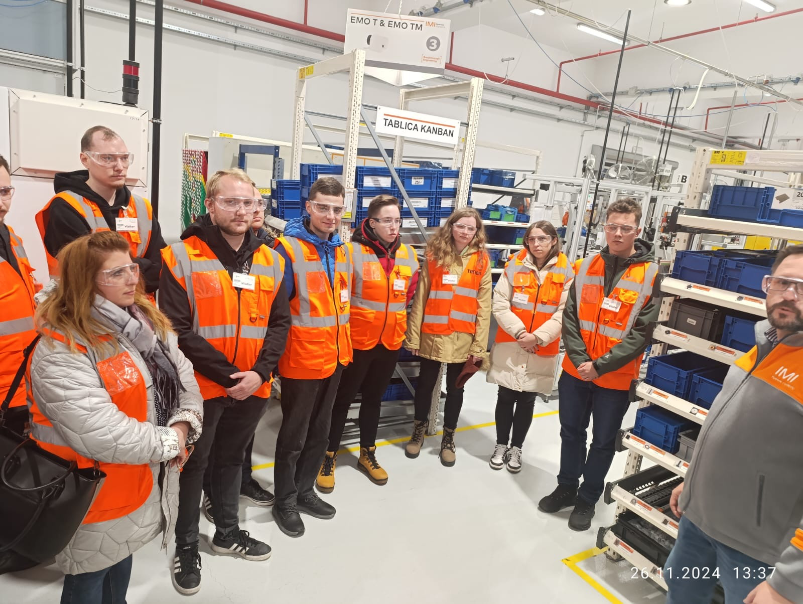 Grupa osób w kamizelkach odblaskowych i okularach ochronnych stoi w hali produkcyjnej.