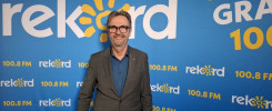 Mężczyzna w garniturze stoi przed niebieskim tłem z logo "Radio Rekord" i napisem "W GRA 100.8 FM".