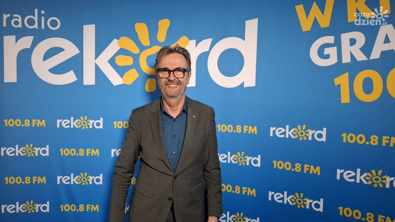 Mężczyzna w garniturze stoi przed niebieskim tłem z logo "Radio Rekord" i napisem "W GRA 100.8 FM".