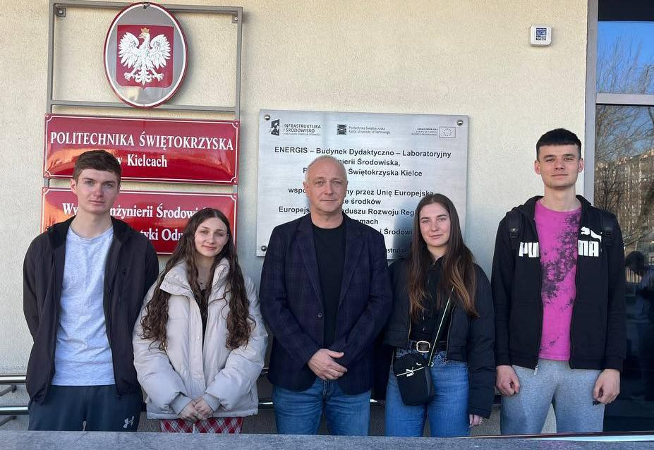 Cztery osoby stoją przed budynkiem Politechniki Świętokrzyskiej. W tle tablice informacyjne i godło Polski.